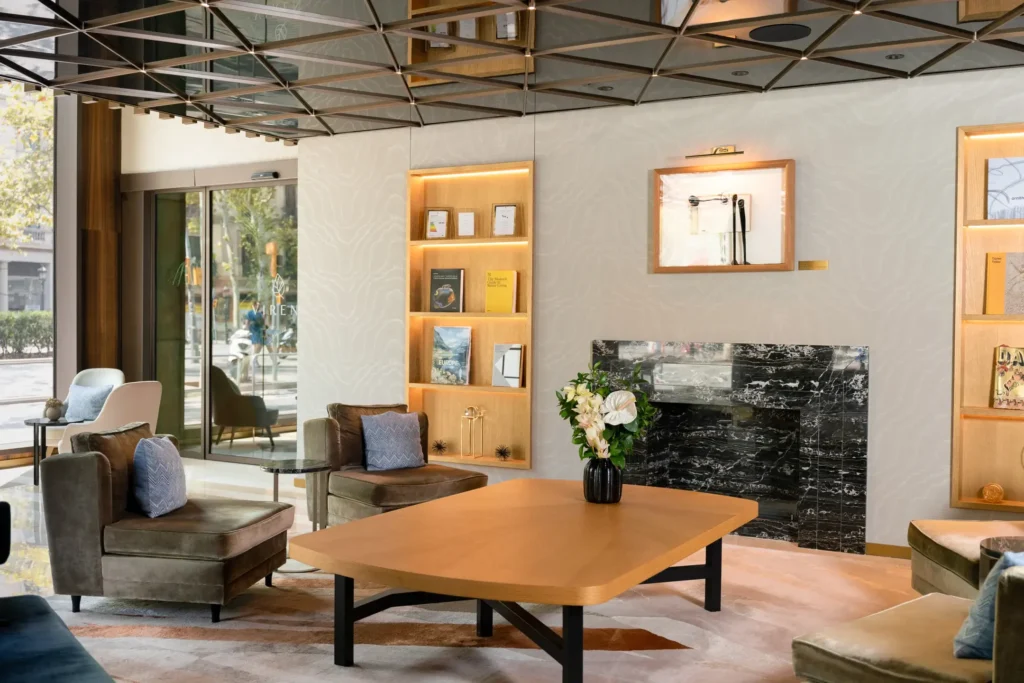 Modern lounge area with plush armchairs, a wooden table, marble fireplace, built-in shelves displaying books and décor, and large windows filling this Inspired Space with natural light.